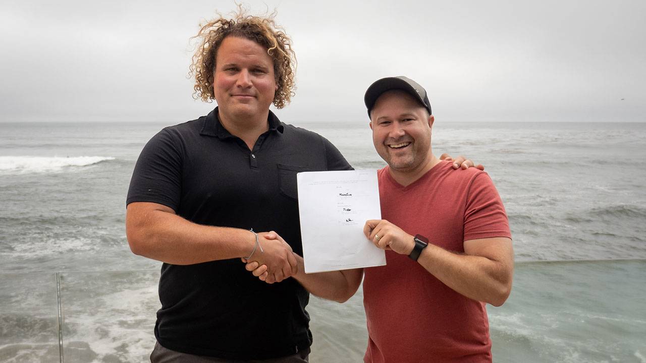 Two men shaking hands and holding signed contract by the ocean on a cloudy day