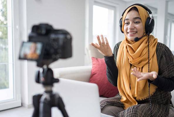 Young woman with headset recording video blog indoors with camera and laptop nearby