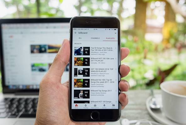 Hand holding smartphone showing Billboard music playlists on YouTube app with laptop and coffee nearby