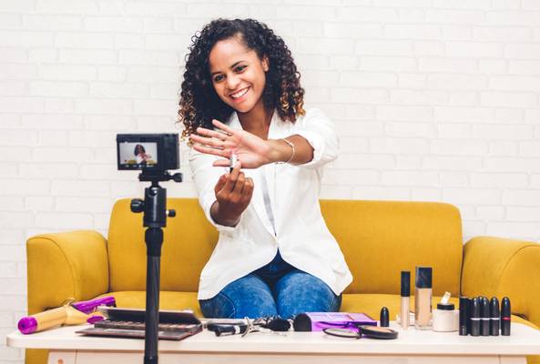 Woman recording beauty vlog showing lipstick on camera from yellow couch with makeup products