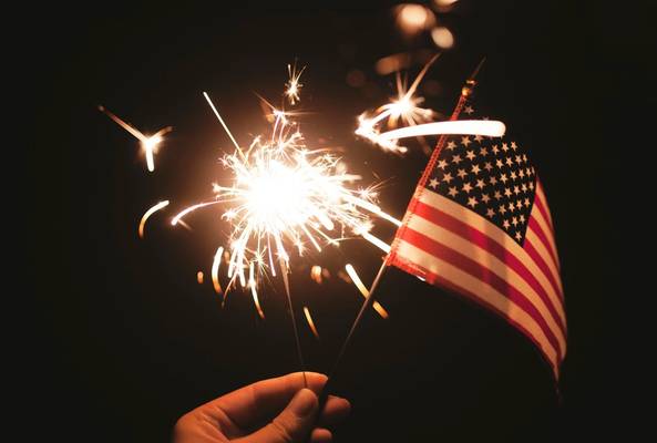 Hand holding lit sparkler and small American flag on dark background celebrating USA.