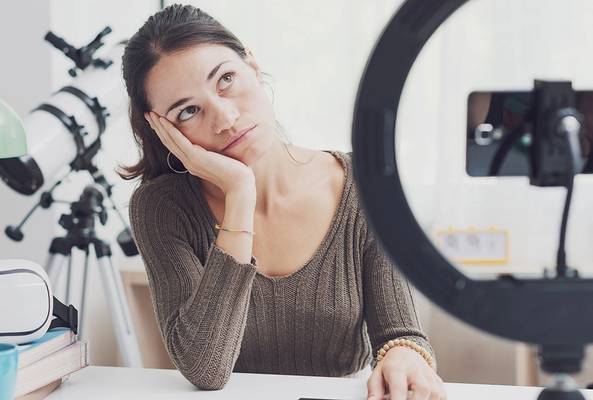 Woman looking bored while recording video with smartphone and ring light at desk