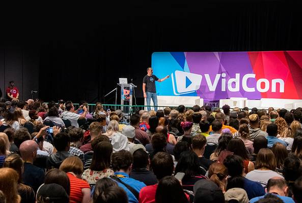 Speaker presenting at VidCon conference with large audience and colorful stage backdrop