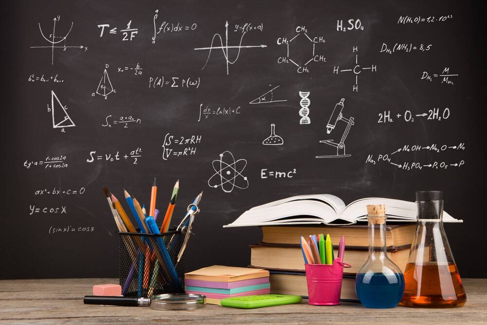 Classroom desk with books, pencils, beakers, and math and chemistry formulas on chalkboard