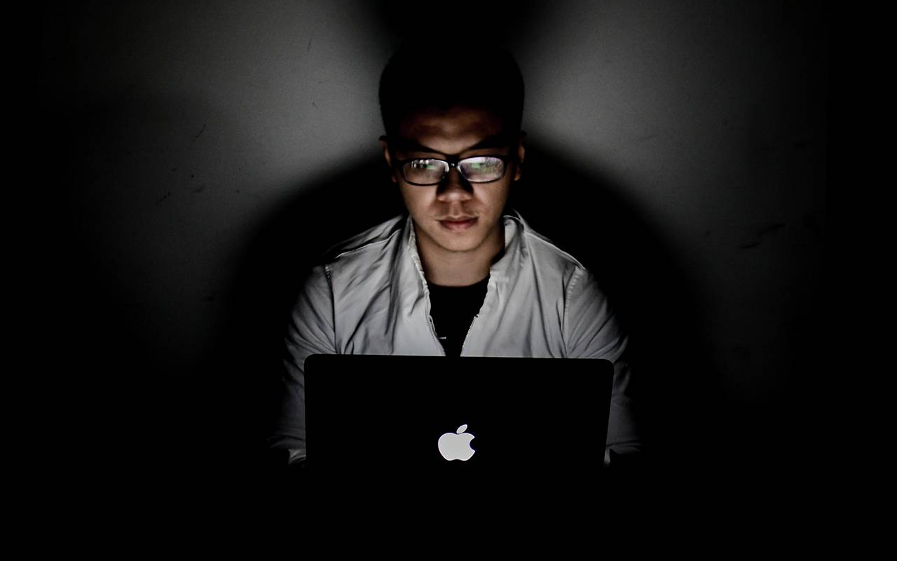 Person wearing glasses working late on a MacBook laptop in a dark room.