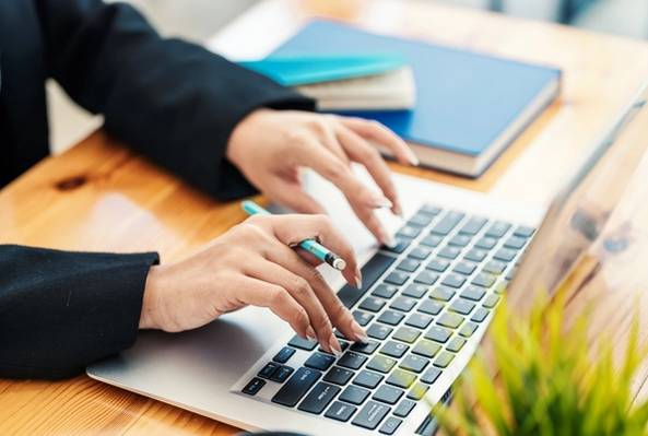 Hands typing on laptop keyboard with pencil, books in background on wooden desk