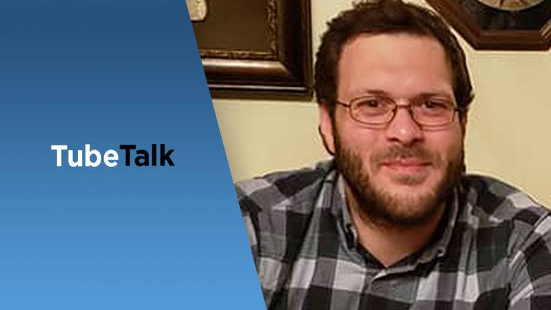 Man in glasses smiling next to blue panel with TubeTalk logo for video podcast.
