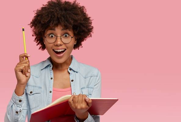 Excited young woman with glasses holding pencil and notebook on pink background