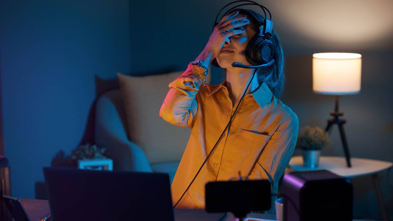 Frustrated gamer wearing headset sitting at desk with laptop and gaming equipment at night