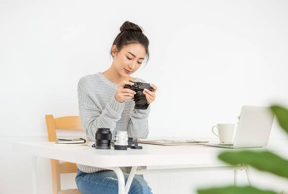 Woman with camera and lenses inspecting photos at a white desk with a laptop and notebook