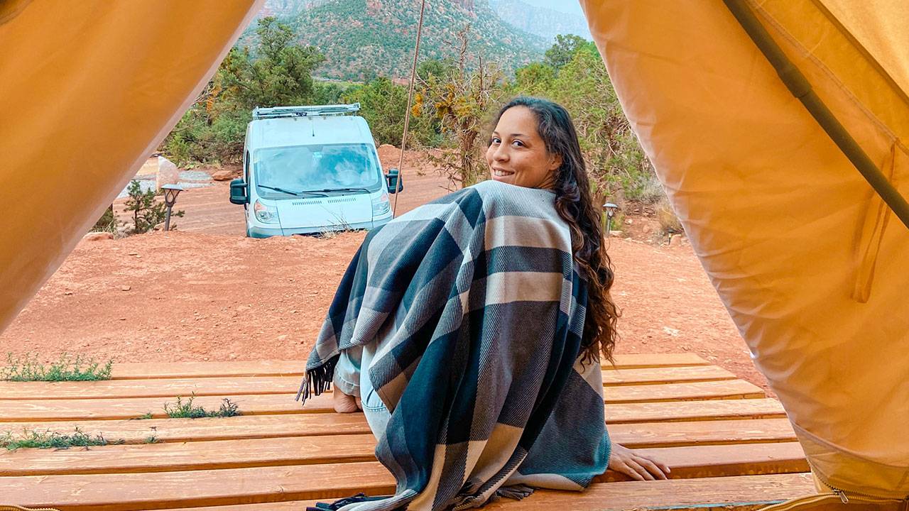 Woman wrapped in a plaid blanket sitting on wooden deck with camper van and trees outdoors
