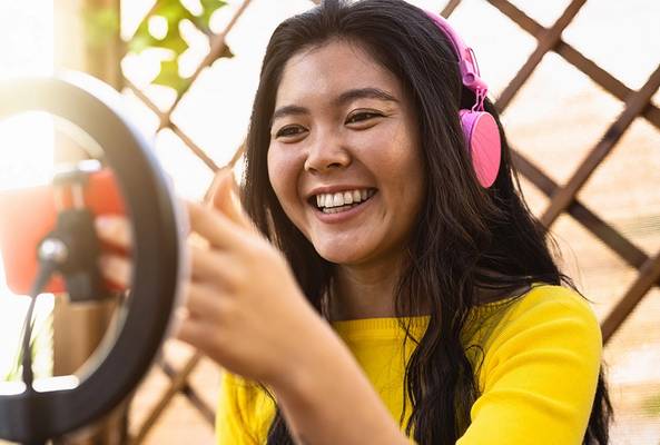 Young woman with pink headphones recording video using smartphone and ring light outdoors
