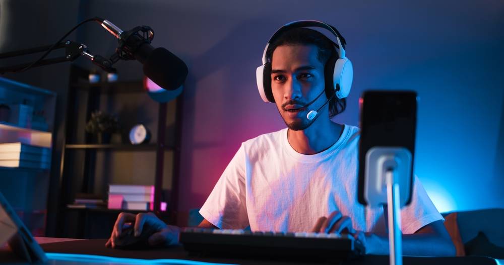 Young male streamer gaming and chatting with headset and microphone in dim room