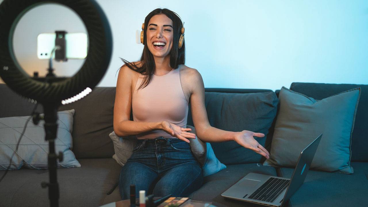 Young woman wearing headphones recording video with ring light and laptop on sofa indoors