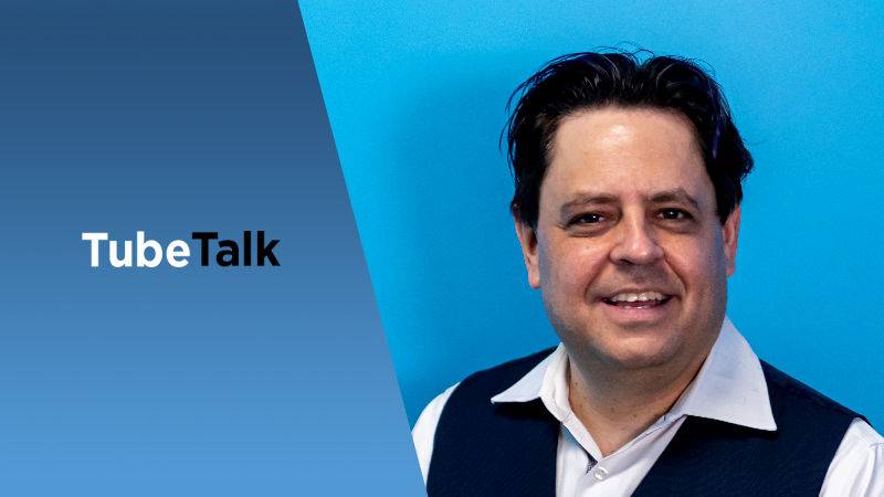 Man in white shirt and vest smiling against blue background with TubeTalk logo