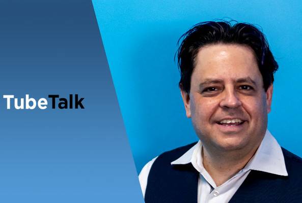 Man in white shirt and vest smiling against blue background with TubeTalk logo