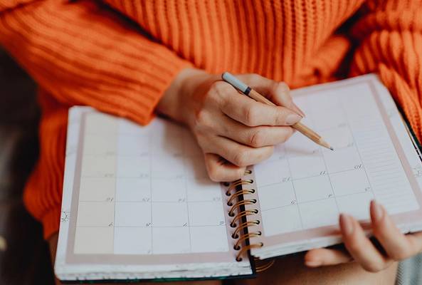 Person in orange sweater writing in a spiral planner calendar with a pencil.