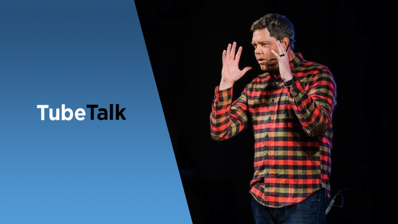 Speaker in red plaid shirt gesturing during TubeTalk presentation on black background