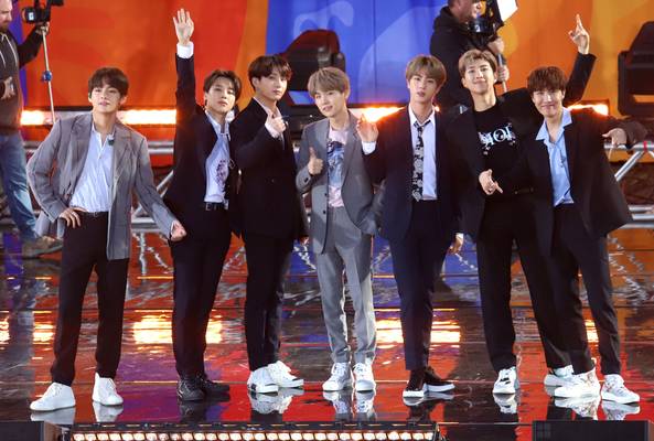 Seven male performers in suits posing on stage with colorful lighting and cameras behind them