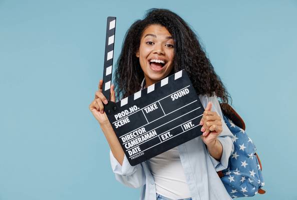 Young woman holding a film clapperboard smiling against blue background with backpack
