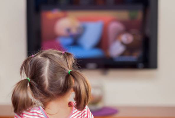 Child with pigtails watching a cartoon on a wall-mounted television in living room.