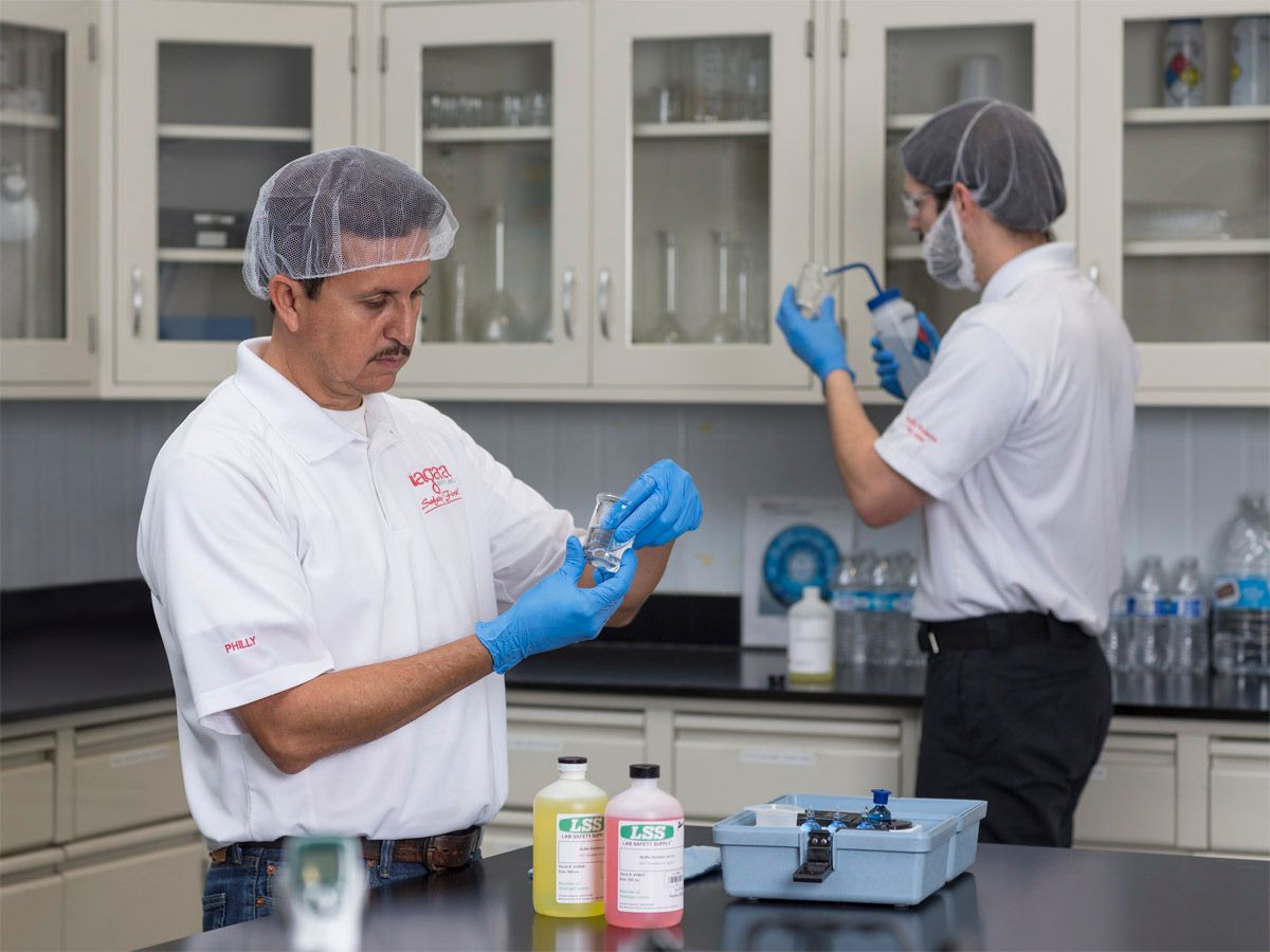 Two Niagara Bottling laboratory employees wearing hairnets and gloves conduct water quality testing in a laboratory setting using sampling equipment and testing materials.