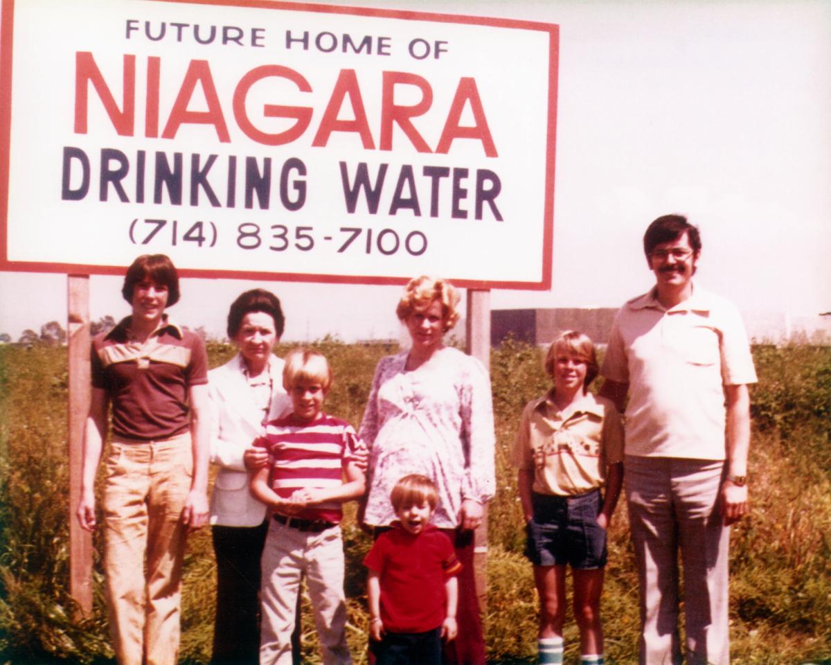 Historic photograph of the Peykoff family standing in front of a sign reading “Future Home of Niagara Drinking Water” with a phone number displayed below.