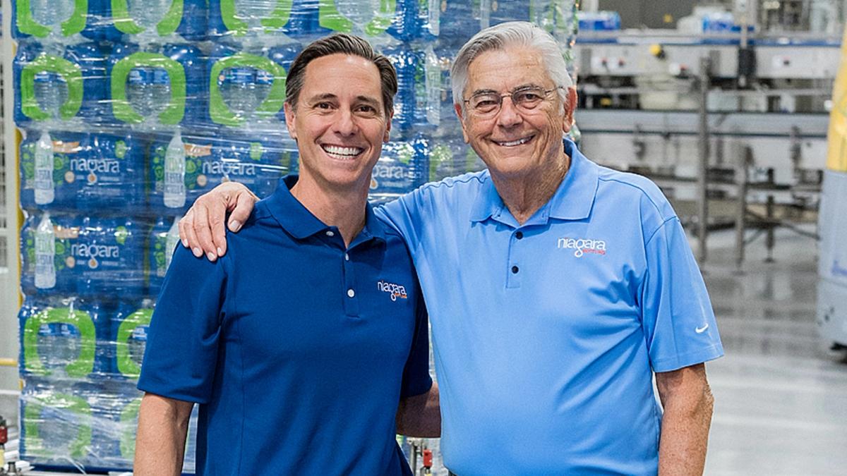 Two individuals wearing Niagara Bottling shirts pose in a beverage manufacturing facility. Pallets of bottled water and production machinery are visible behind them.