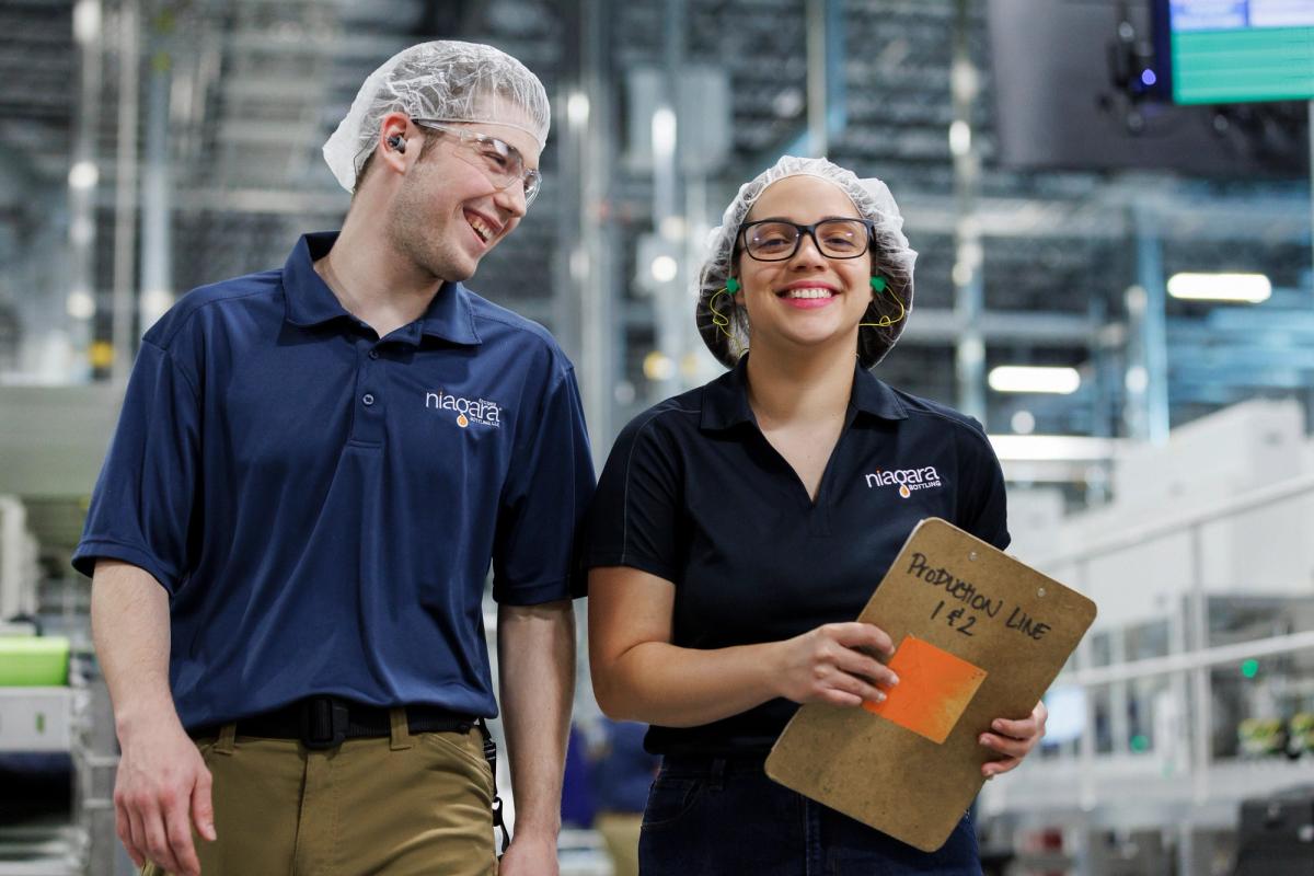 Two employees wearing hairnets, safety glasses, and hearing protection stand next to a bottled water production line, with manufacturing equipment visible throughout the facility.