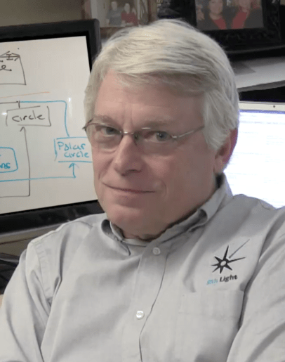 Man with white hair and glasses wearing light gray shirt with logo, sitting at desk with whiteboard diagram visible behind him.