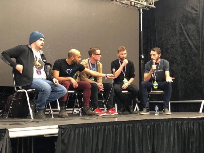 Five men sit on stage during a panel discussion, with one speaking into a microphone while others listen and engage.