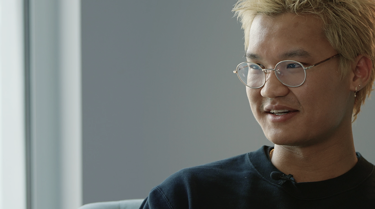 Person with blonde spiky hair and round glasses wearing a black shirt, smiling indoors.