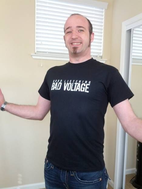Man wearing black "Bad Voltage" t-shirt standing in front of white blinds with arms outstretched.