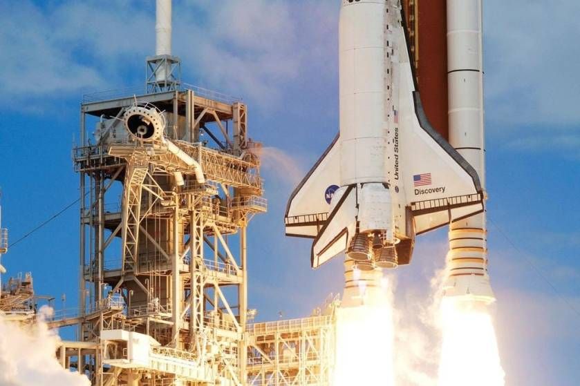 Space Shuttle Discovery launching from its launch pad with engines firing and support tower visible against blue sky.