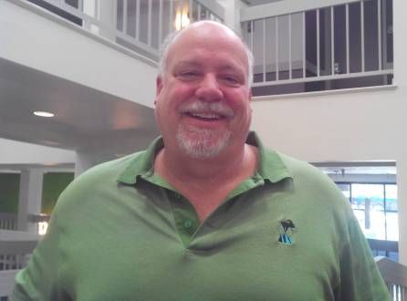 Smiling man in green polo shirt standing in a modern indoor venue with white walls and railings.