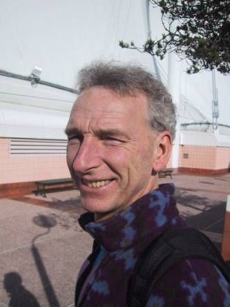 Middle-aged man smiling at camera outdoors with industrial building in background.