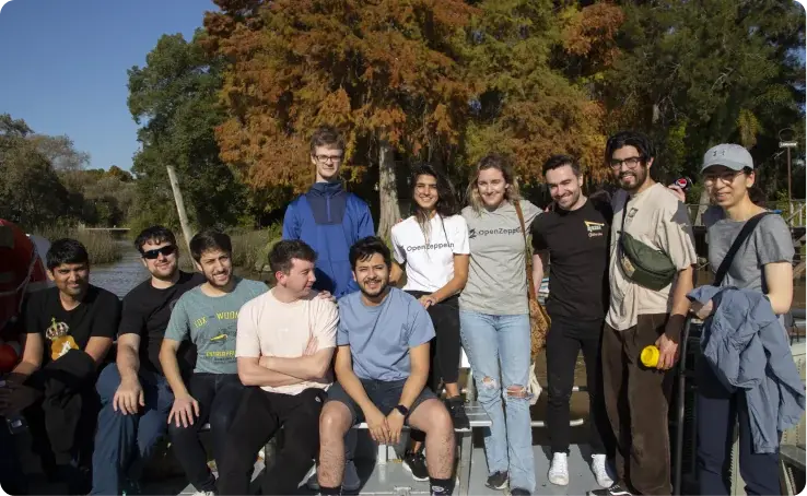 Group of eleven people posing together outdoors by a waterway with autumn trees in the background.