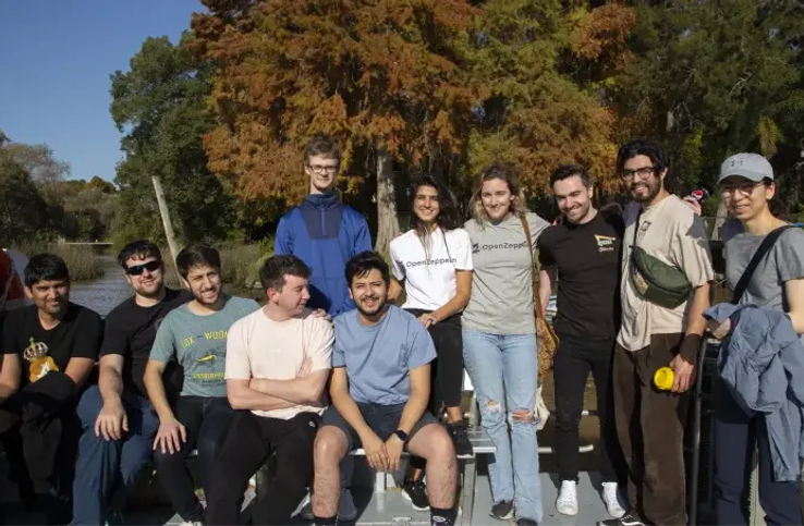 Group of eleven people posing together outdoors by a waterway with autumn trees in the background.