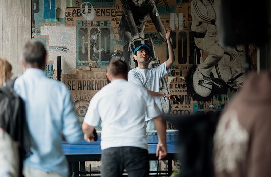 Man in white shirt reaching up toward skateboard mounted on wall covered with vintage signage while others watch nearby.