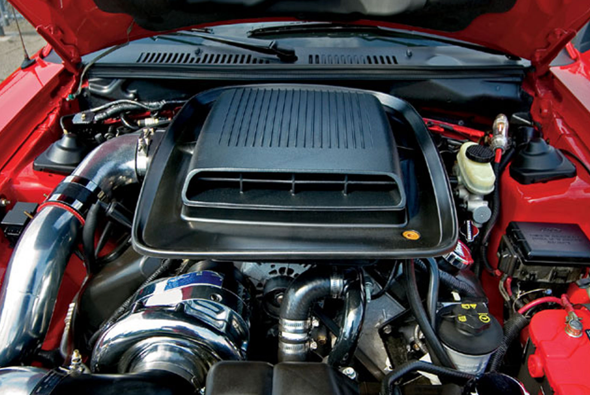 Red muscle car engine bay with black hood scoop and polished chrome supercharger components.