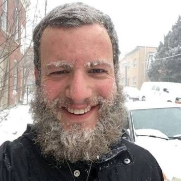 Man with full beard smiling outdoors in snowy winter setting near parked vehicles.