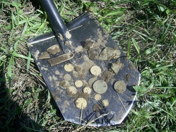 Shovel covered in soil with various coins scattered on the blade, resting on grass and dirt.