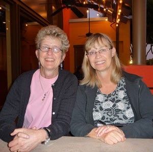 Two women smiling at the camera while seated at a table in an indoor venue with warm lighting.