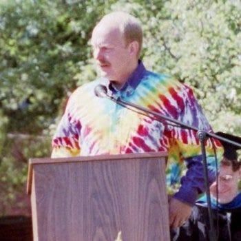 Man in tie-dye shirt speaking at wooden podium with microphone outdoors.