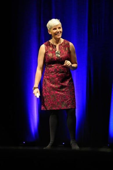 Woman with short blonde hair speaking on stage wearing a red patterned sleeveless dress against a blue-lit backdrop.