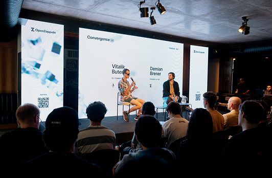 Two speakers sit on stage during a conference panel discussion while audience members watch from their seats.