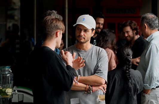 Man in white cap and gray shirt conversing with others at a social gathering indoors.