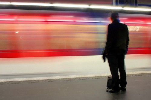 Person in dark clothing standing on platform as red and white train passes by in motion blur.