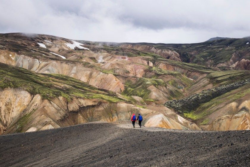 Two hikers in colorful jackets stand on a dark volcanic plateau overlooking a vast valley with multicolored mineral-striped mountains and green slopes.