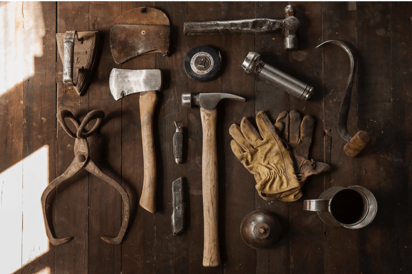 Collection of vintage hand tools arranged on dark wood surface including hammers, axes, pliers, wrenches, leather gloves and metal hardware.